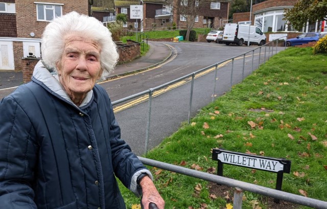 an older woman standing in front of a street sign labelled Willett Way and holding a walking stick as she smiles at the camera