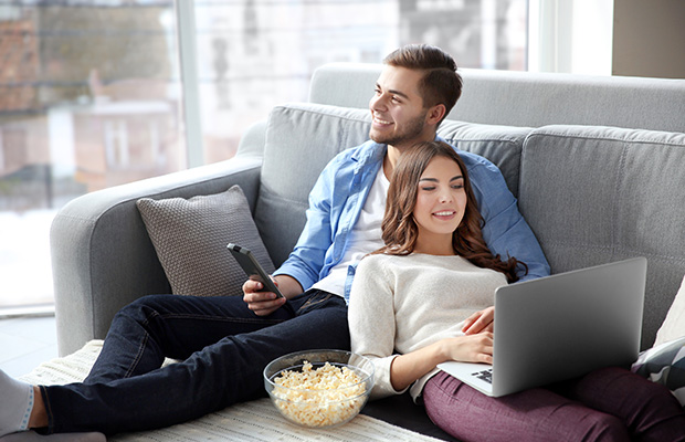A couple sitting down on the sofa watching TV and looking at a laptop