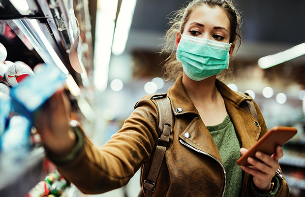 A person wearing a face mask holds a mobile phone in one hand and takes a packet from a supermarket shelf with the other