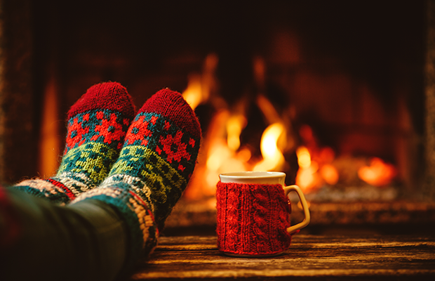 A pair of feet in thick patterned socks in front of an open fire next to a mug of tea with a knitted red jacket.