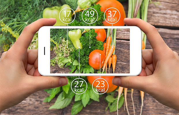 two hands hold a smartphone over some vegetables, the screen shows how many calories they have