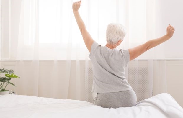 A grey-haired woman sits on a bed doing a morning stretch.