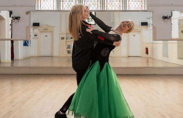 Katharine Holmes wears a green and black ball gown and dances with her teacher in large dance hall.
