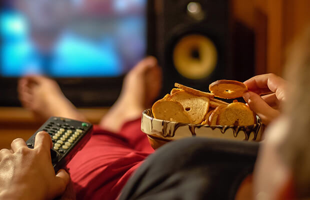 A person watches TV with a remote control in their left hand and their right hand in a bowl of snacks