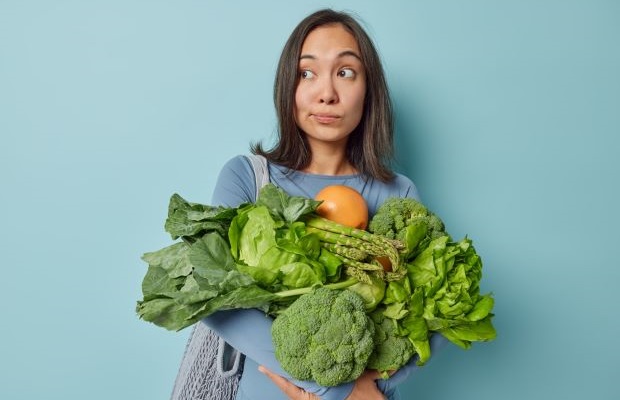 A person holds a variety of green vegetables with a quizzical expression on their face.