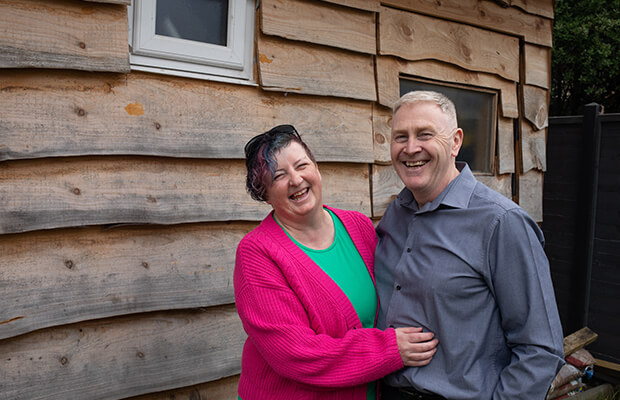 Maggie Sewell laughs with her hands around her husband Rob in front of a wooden building