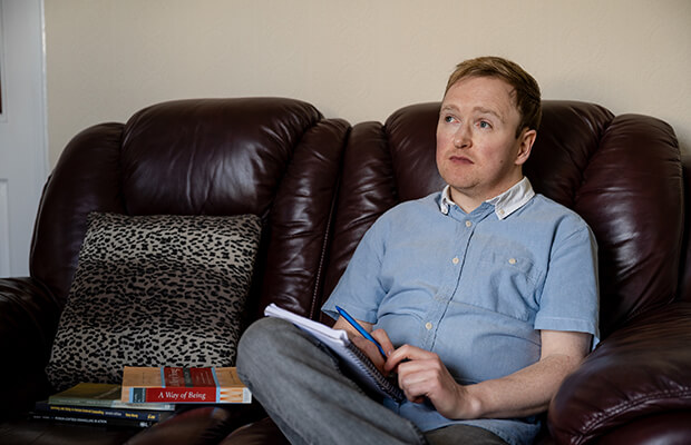 Patrick McCann sits on a leather sofa with a notebook and pen in his hand