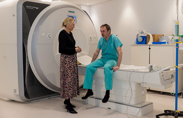 An adult sits on an MRI scanner table and talks to Charlotte Manisty who is standing next to them.