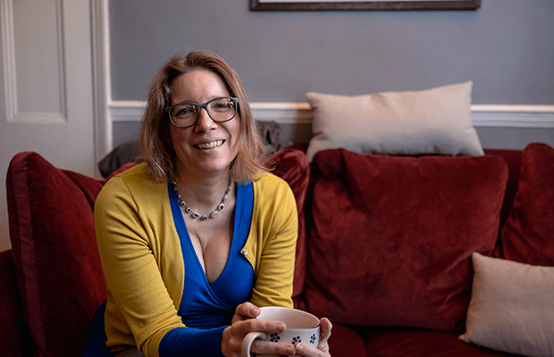Kerstin Sailer sits on a red velvet sofa with a cup in her hands and smiling