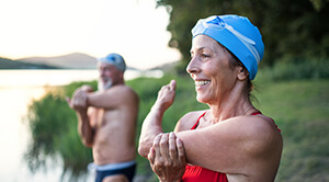 Two swimmers with plastic caps and swimming goggles stretch their arms next to a lake