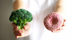A person holding a broccoli in their right hand and a pink iced donought in their left hand