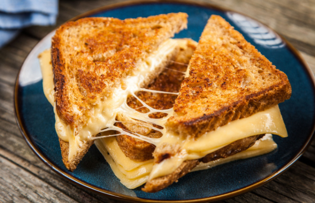 A cheese toastie made of white bread cut in half with melted cheese hanging from it, sat on top of an uncut cheese toastie. 