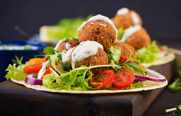 An open, white wrap on a chopping block with lettuce, tomato, onion, falafel and mint yoghurt on top ready to be wrapped up. 
