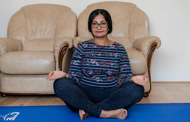 Hina Shah sits crossed legged on a blue yoga mat in Sukhasana easy pose