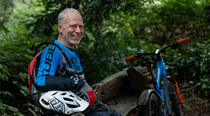 Neil Caton sitting on a log in a forest next to his electric bike.