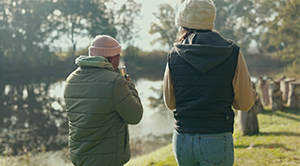Two people in coats and hats walking beside a river