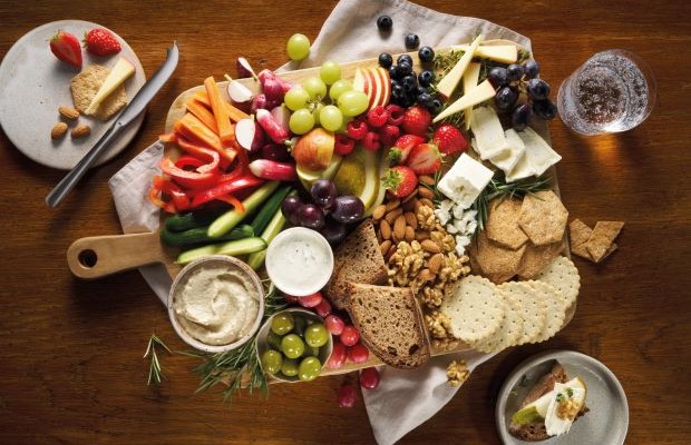 Arial view of a healthy cheese board with sliced vegetables, fruit, nuts, crackers and sliced cheese.