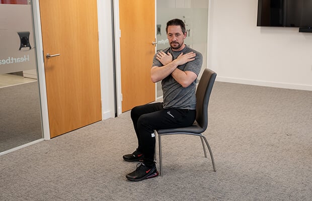 Andrew Scard demonstrates seated trunk rotation on a chair with his arms crossed on his chest as he turns to his left