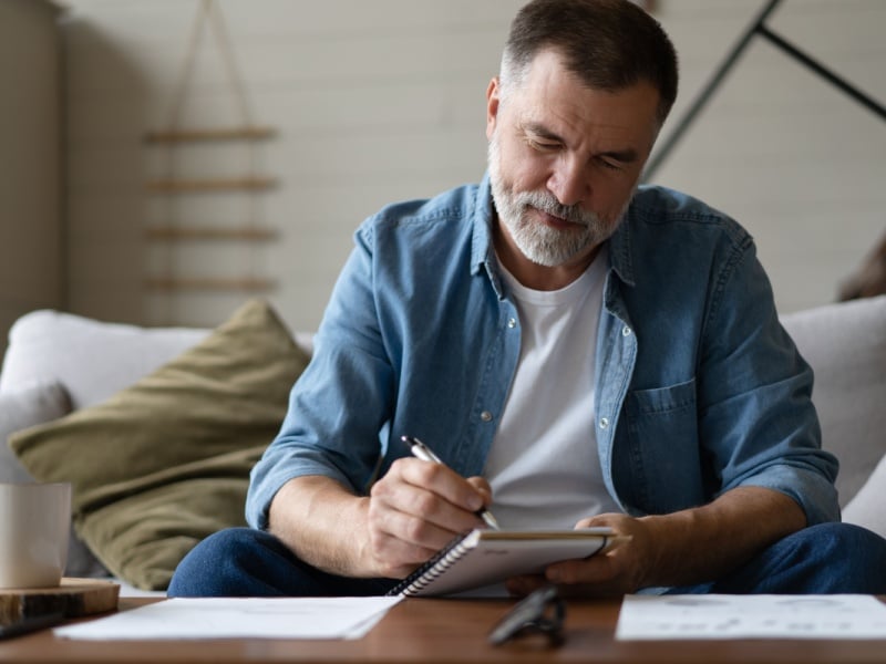 A man fills out a diary while sitting on his sofa.