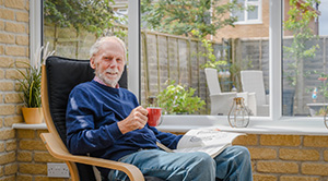 Glynn Evans sitting in a chair in his home with a book and a mug.