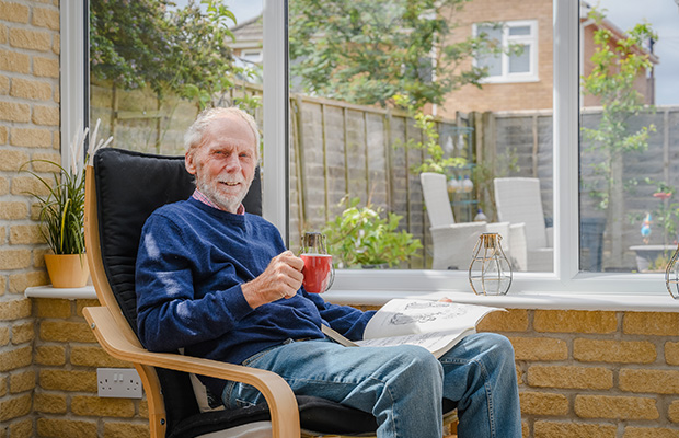Glynn Evans sitting in a chair in his home with a book and a mug.