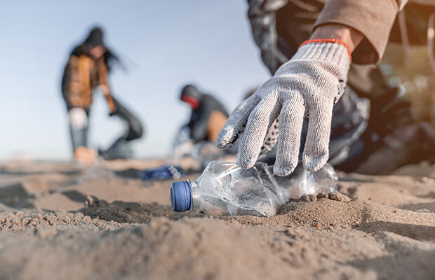 A person with a gloved hand reaches to pick up a plastic bottle on a beach with other litter pickers in the distance