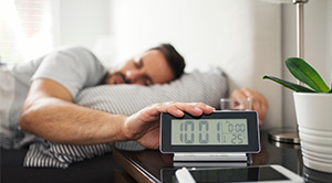 A man in bed in the morning reaches for his alarm clock.