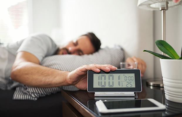 A man in bed in the morning reaches for his alarm clock.