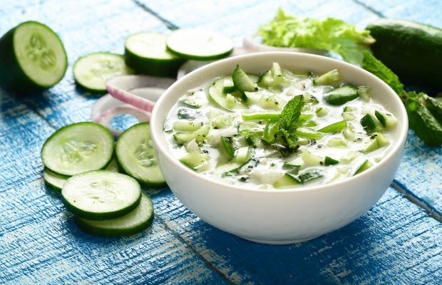A bowl of cucumber and yogurt dip with fresh herbs on a blue table.
