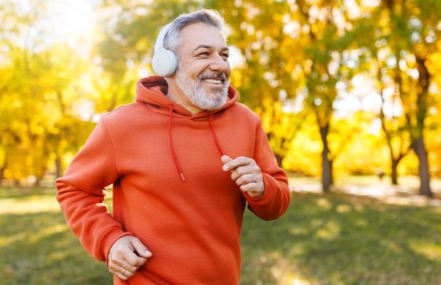 A middle-aged man wearing an orange hoodie and headphones running through a park, smiling.