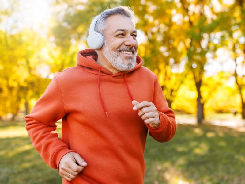 A middle aged man wearing an orange hoodie and headphones running through a park, smiling.