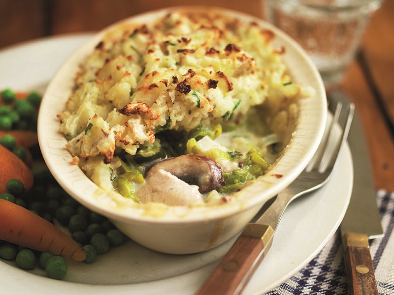 Close up of an individual pie dish containing chicken, leek and mushroom pie with a crispy top and sitting on a plate alongside some peas and carrots.