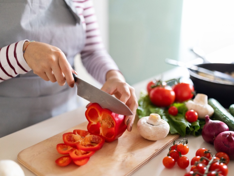 Close up of a person wearing a red and white striped top chopping a red bell pepper on a chopping board with a large knife.