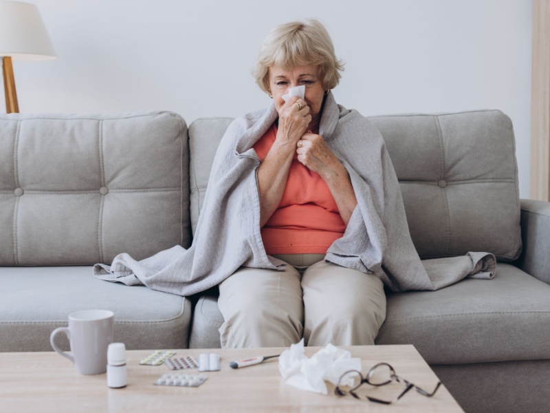 A senior woman wearing a orange top and beige trousers sits on a grey sofa with a grey blanked around her shoulders, blowing her nose into a tissue. A cup and some used tissues on the coffee table in the foreground.