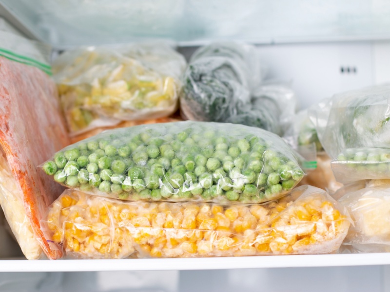 Frozen vegetables, including peas and sweetcorn, in clear plastic pouches on a shelf in a freezer.