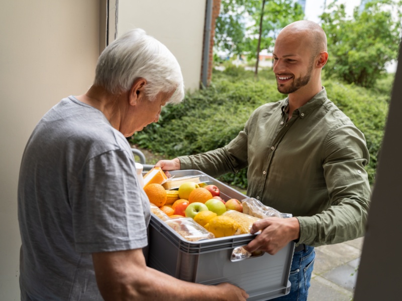 A supermarket delivery driver in a green shirt stands in a doorway, delivering a box of groceries to a grey-haired man in a grey t-shirt.