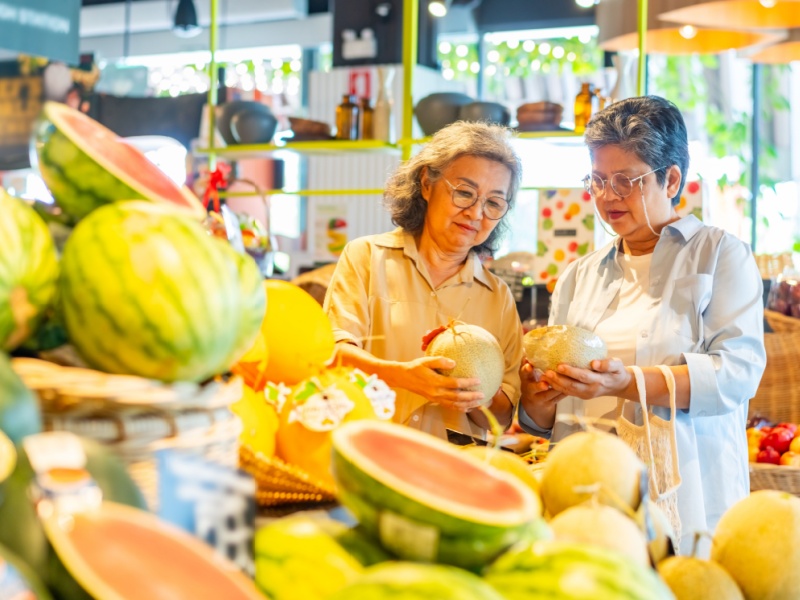In a supermarket with a display of watermelons in the foreground and two senior women choosing some fruit in the background.