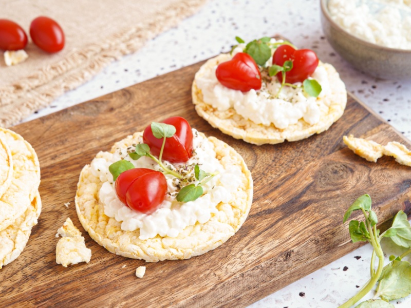 Two rice cakes with cottage cheese and halved cherry tomatoes, on a wooden board.