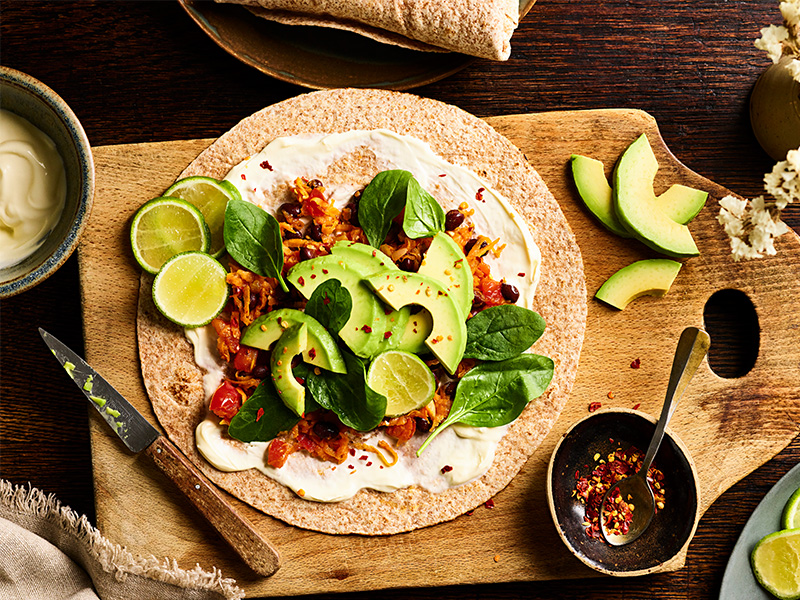 An open tortilla wrap with black beans, avocado, sweet potato and spinach leaves.