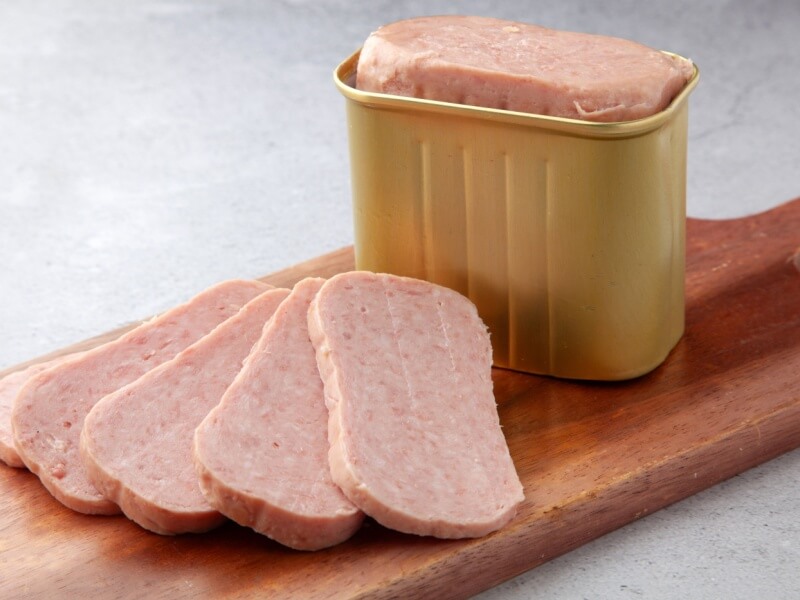 Slices of canned meat in a tin and on a wooden board.