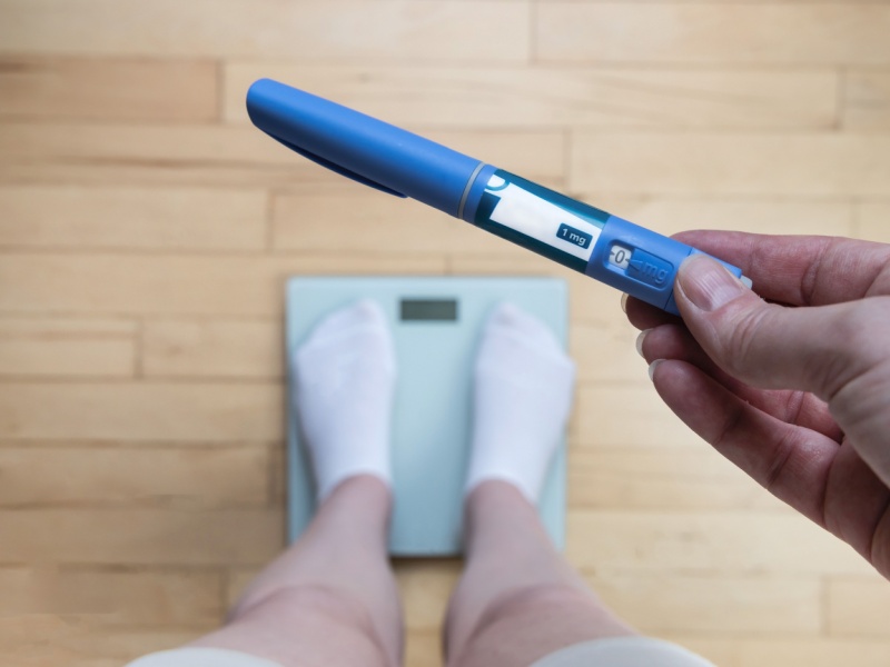 A person standing on scales holding a weight-loss injection pen.