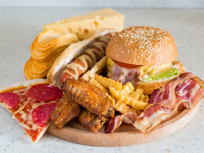 A wooden board filled with bacon, pizza, chicken wings, cheese, chips, a hot dog and burger, illustrating the types of food to avoid if you have high cholesterol.