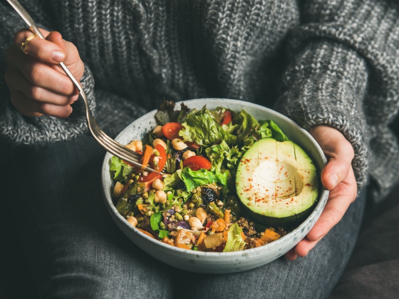 Woman's hands in a warm grey jumper holding a healthy vegan meal with avocado, vegetables and pulses