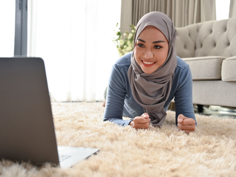 Muslim girl wearing a hijab doing exercises at home on the floor and looking at a laptop.