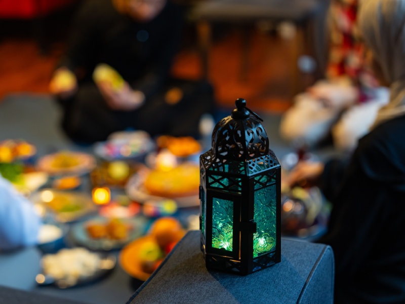 A Muslim family sit around a table with plates of food in the background during iftar, a lantern is in the foreground.