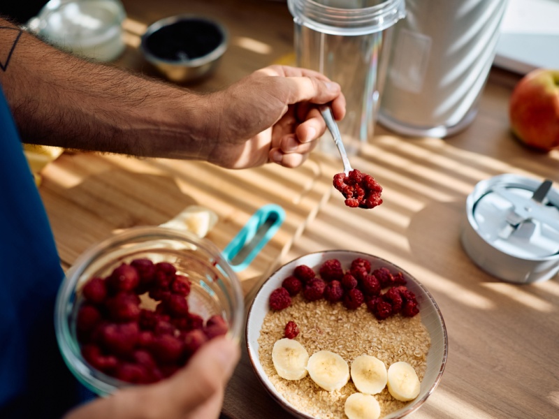 Close up of a man's hand spooning raspberries onto a bowl of porridge with slices of banana, on a wooden worktop.
