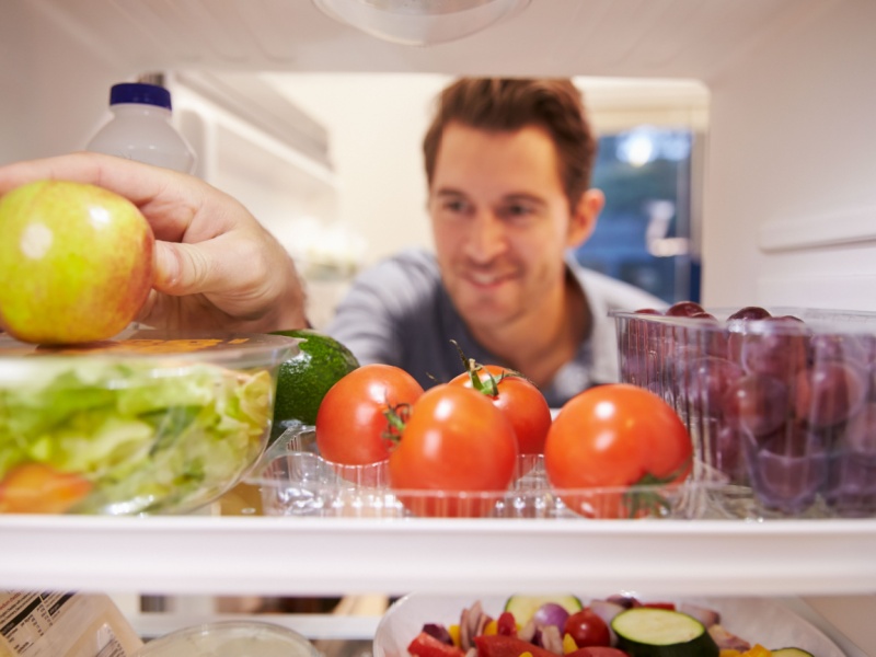 View from inside a fridge showing a man opening the fridge door and picking an apple from the top shelf, which is next to some tomatoes and grapes.