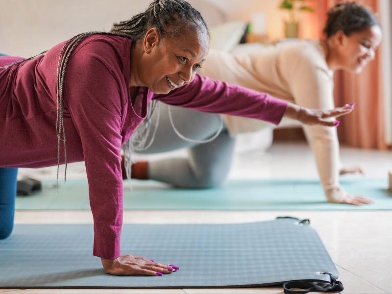 A senior woman of diverse origin wearing a purple top doing exercises on a mat, with her daughter next to her.