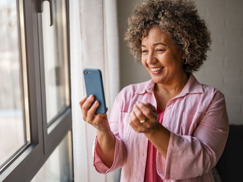 A mixed race woman wearing a pink shirt smiles as she holds her phone up to her face during a video call.
