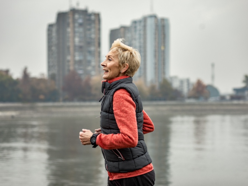 A senior woman with short blonde hair wearing a red fleece under a black gilet, running on a grey winter day next to a river and some tower blocks.